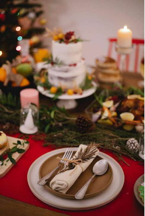 Gedekte kersttafel versiert met dennentakken, dennenappels en kaarsen met kerstboom op de achtergrond.