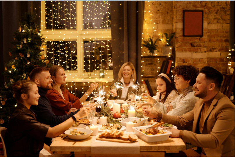 7 mensen zitten rond een tafel met eten en vieren Kerstmis in een versierde kamer met veel lichten.