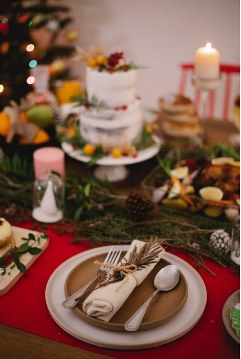 Set Christmas table adorned with pine branches, pinecones, and candles with a Christmas tree in the background.